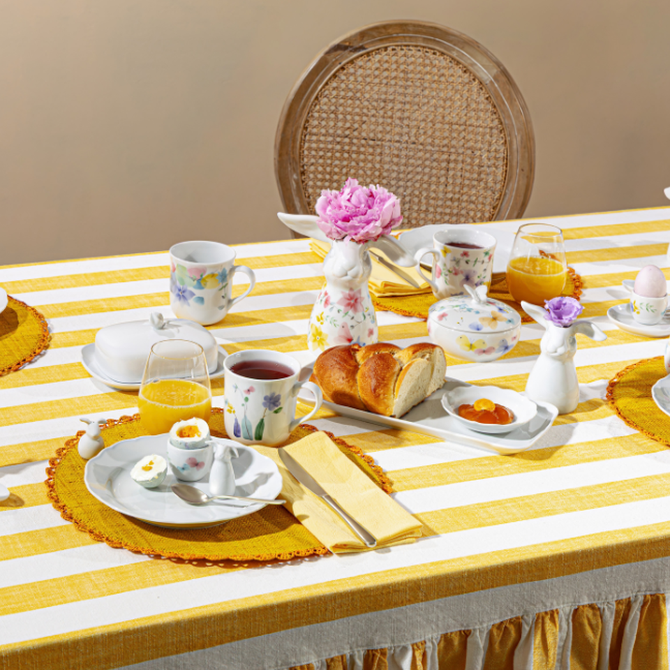 Servizio da tavola pasquale Hutschenreuther con decorazioni floreali su tavola a strisce gialle e bianche, decorato con tazze, piatti, fiori e accenti primaverili.
