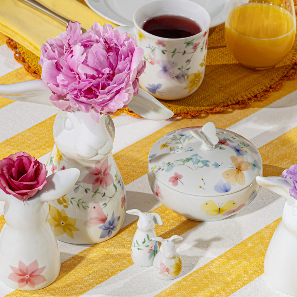 Servizio di piatti colorati Hutschenreuther con un delicato motivo floreale su una tovaglia a righe gialle e bianche, impreziosito da un vaso, una zuccheriera, una tazza e fiori freschi.