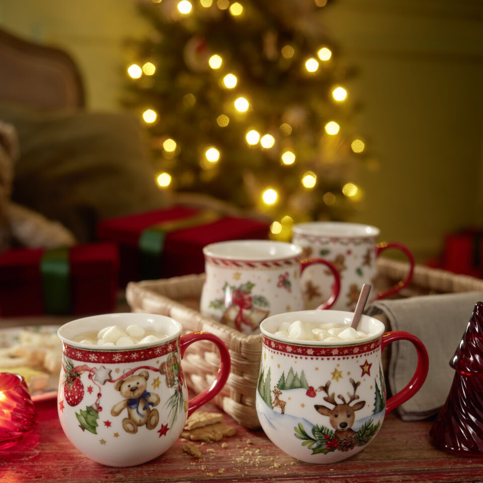 Four mugs with handles with Christmas motifs, filled with a hot drink with small marshmallows visible on the surface of the drink