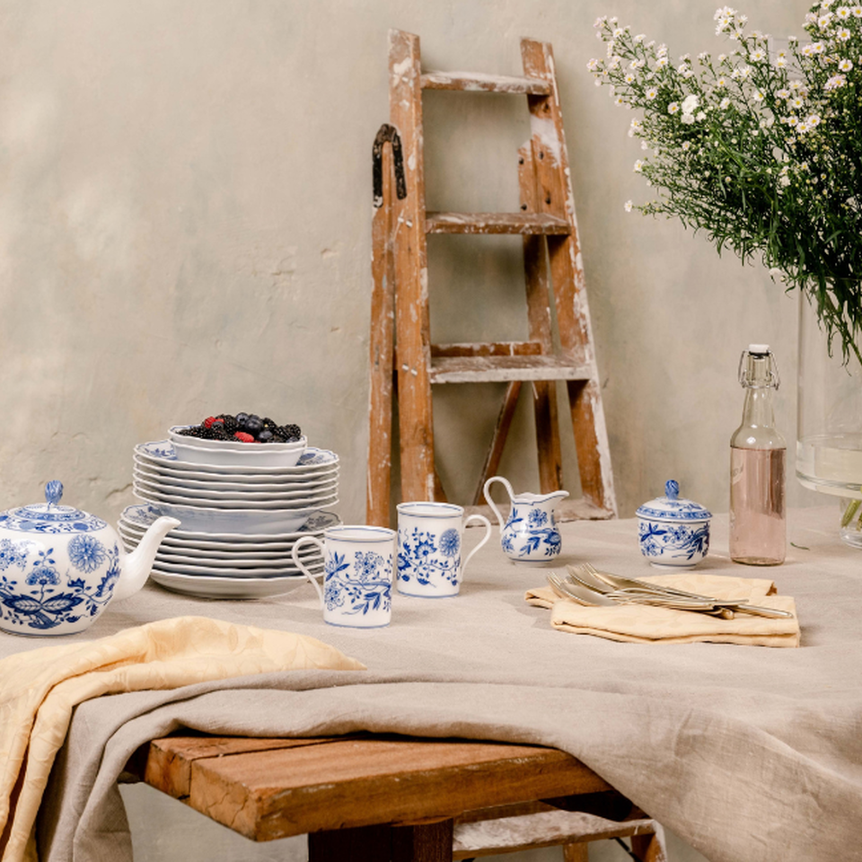 Servizio di porcellana Hutschenreuther blu e bianco con motivi floreali su una tavola elegantemente apparecchiata con piatti, teiera, tazze e fiori freschi sullo sfondo.