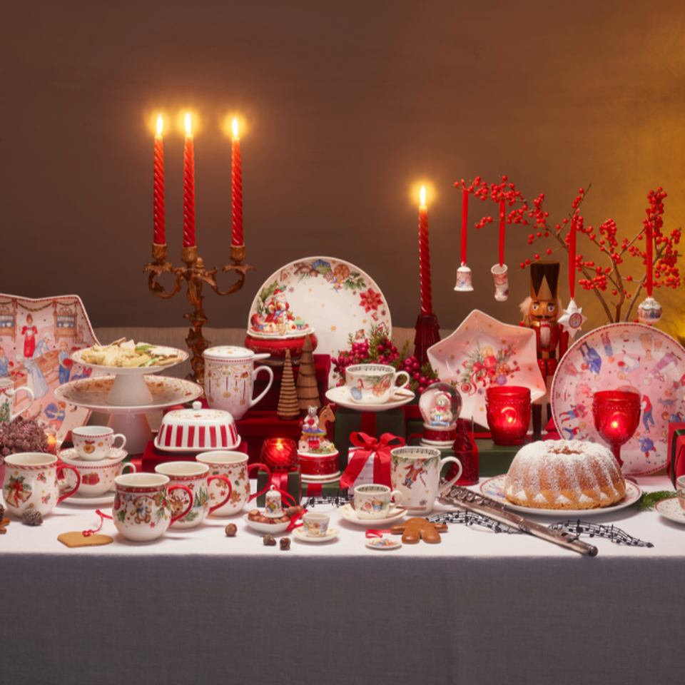 Una tavola imbandita a festa con una tovaglia bianca, stoviglie natalizie rosse e bianche, candele accese e un Gugelhupf, accanto a un albero di Natale illuminato.