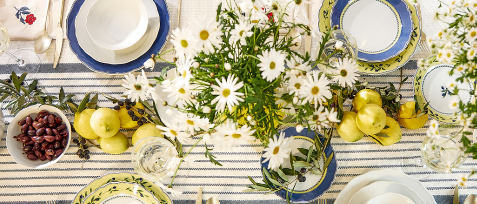 A Mediterranean-style table setting with crockery in blue-and-white and green-patterned designs, decorated with bouquets of daisies, fresh lemons and a bowl of olives.