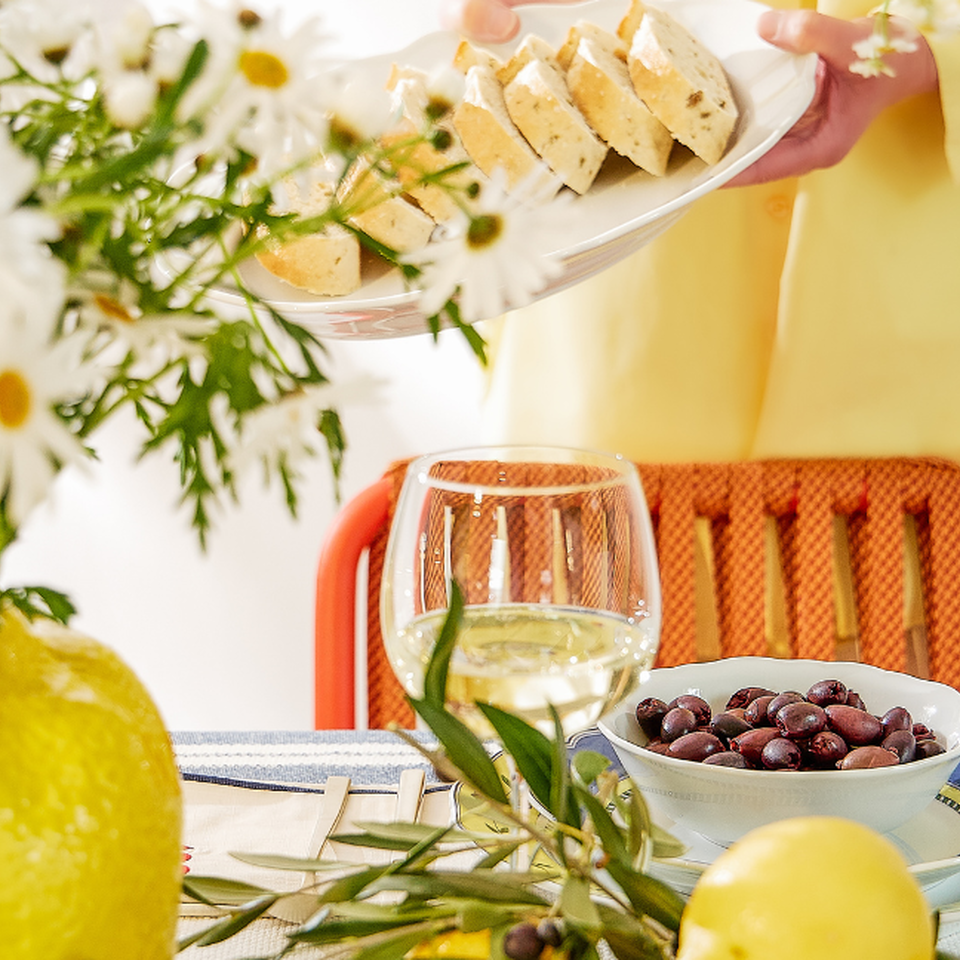 A laid table with a white wine glass, a bowl of olives on blue-and-yellow Hutschenreuther tableware and lemons; in the background, cheese canap&eacute;s are being served.