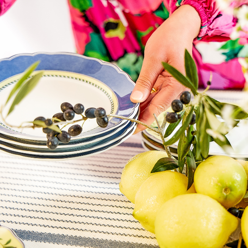 A person in a floral blouse is holding a stack of blue and white Hutschenreuther plates with an olive branch; in front of them are lemons on a striped table runner