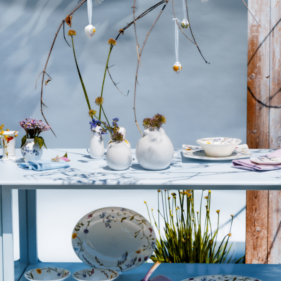 Servizio di piatti Hutschenreuther con motivi floreali su una mensola decorativa all'aperto, con piatti, ciotole, vasi e rami primaverili disposti in un elegante allestimento scenografico.