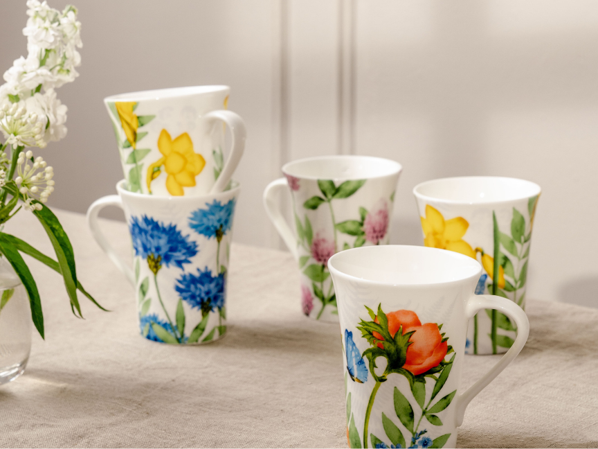 Hutschenreuther mug with colourful floral pattern, arranged on light-coloured fabric next to a vase with white flowers.
