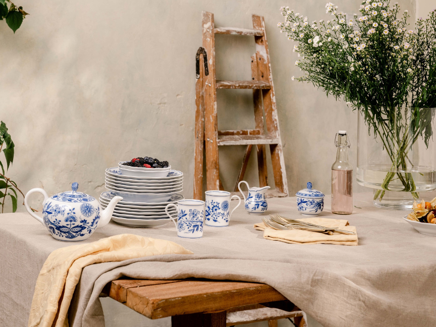 Hutschenreuther blue and white porcelain tableware with floral pattern on an elegantly laid table with plates, teapot, cups and fresh flowers in the background.