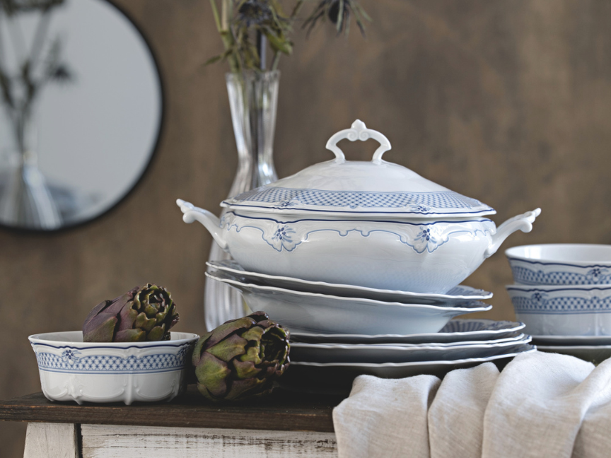 Blue and white porcelain tableware from Hutschenreuther with delicate border decoration, consisting of a soup tureen with lid, deep and shallow plates, and bowls, arranged as a classic dinner service on a rustic table.